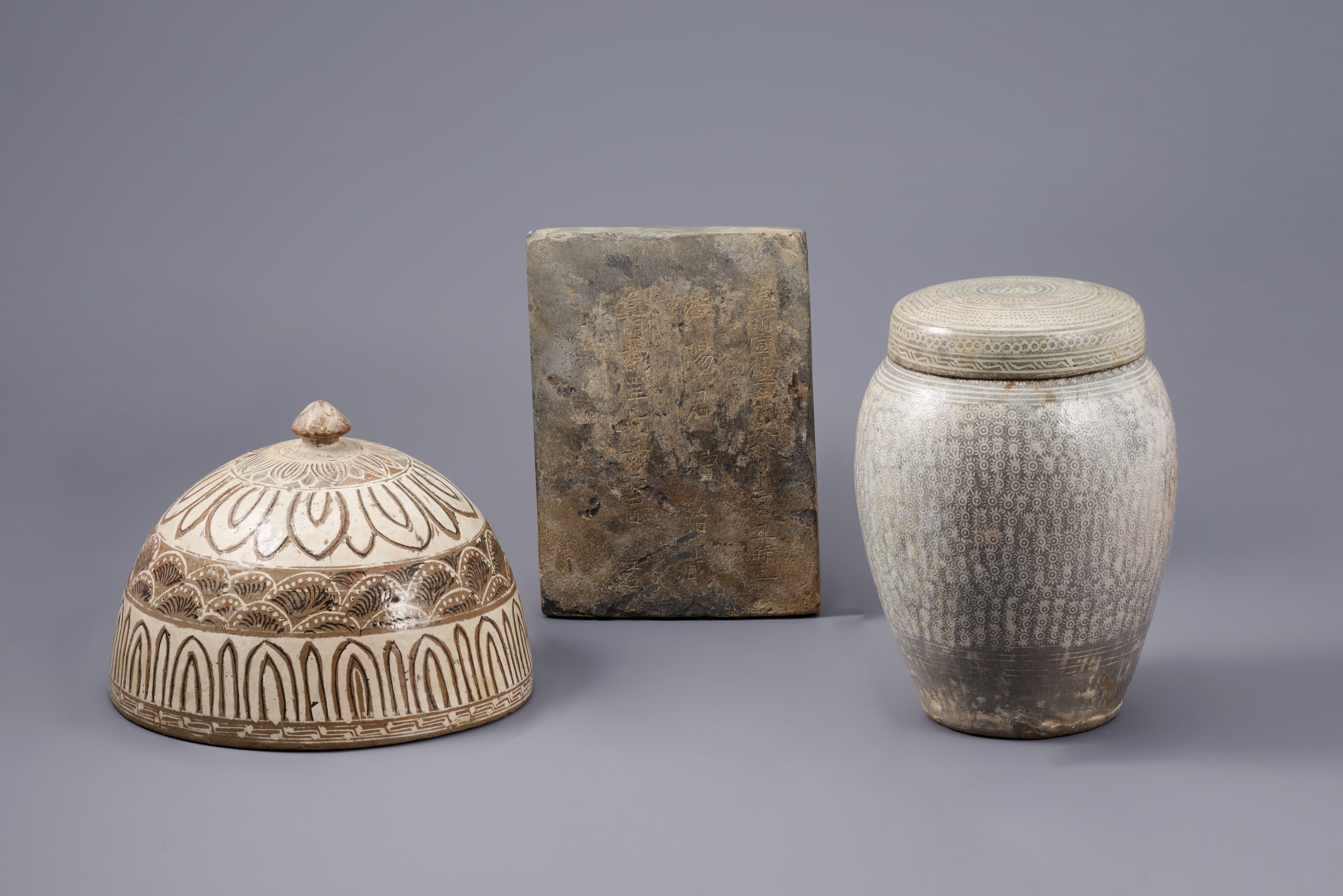 At left sits a half-dome-shaped ceramic, cream-colored with curvilinear patterns in thin brown lines; at center is a vertical brown slab, scratched with vertical lines of calligraphy; and at right is a slightly bulging jar with a dense pattern of circles, dots, and lines in white against a dark background.