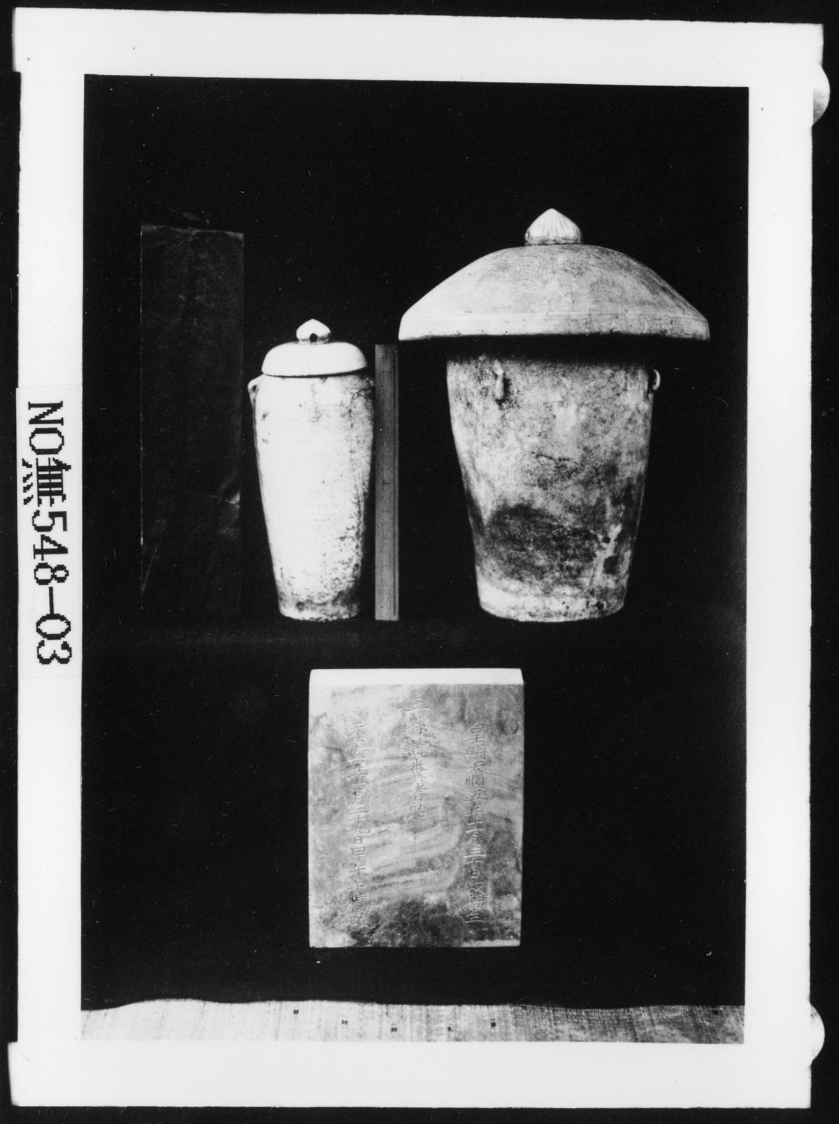 Black and white image of a two ceramic vessels above a mottled rectangular tablet.