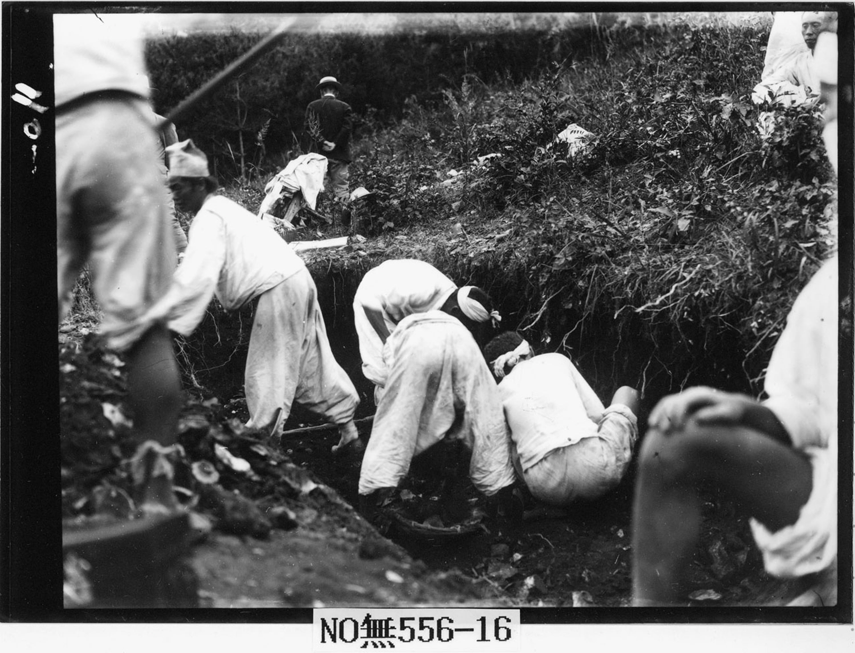 This black-and-white photograph features at least nine people in white shirts and loose pants. In the center, four men sit, crouch, and bend over a hole in the side of a hill. In the middle ground, a man in seemingly western clothes faces away from the camera.