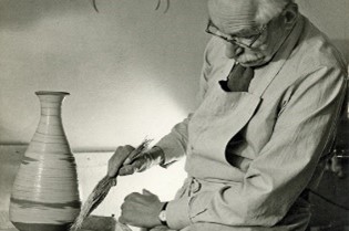 In this black-and-white photograph, a mustached man sits at right and peers down to the left as he uses a brush on a light-colored jar or vase. He wears an apron and glasses.