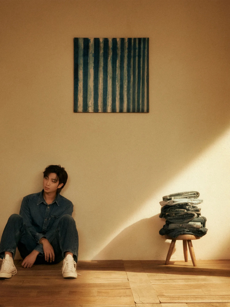 In a shallow space, a person wearing denim and white sneakers sits at the left on a wood floor. At right, a low stool holds a stack of denim clothes, lit by raking light. At upper center is a square artwork featuring thick, dark stripes that seem to bleed into a light background.