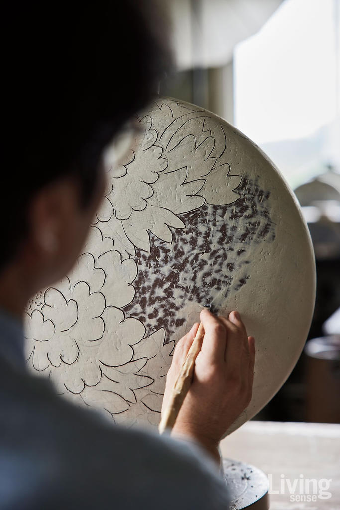 In this photograph, you see over a person's right shoulder as they work on a design on a large, white circular dish with a flower motif on it.