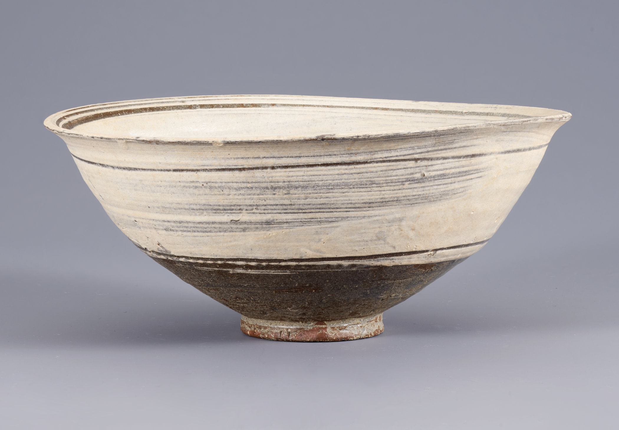 This widely flared bowl sits on a thin base. Not perfectly round or smooth, the bowl features cream-colored slip in the upper two thirds, brushed over the brown substrate.