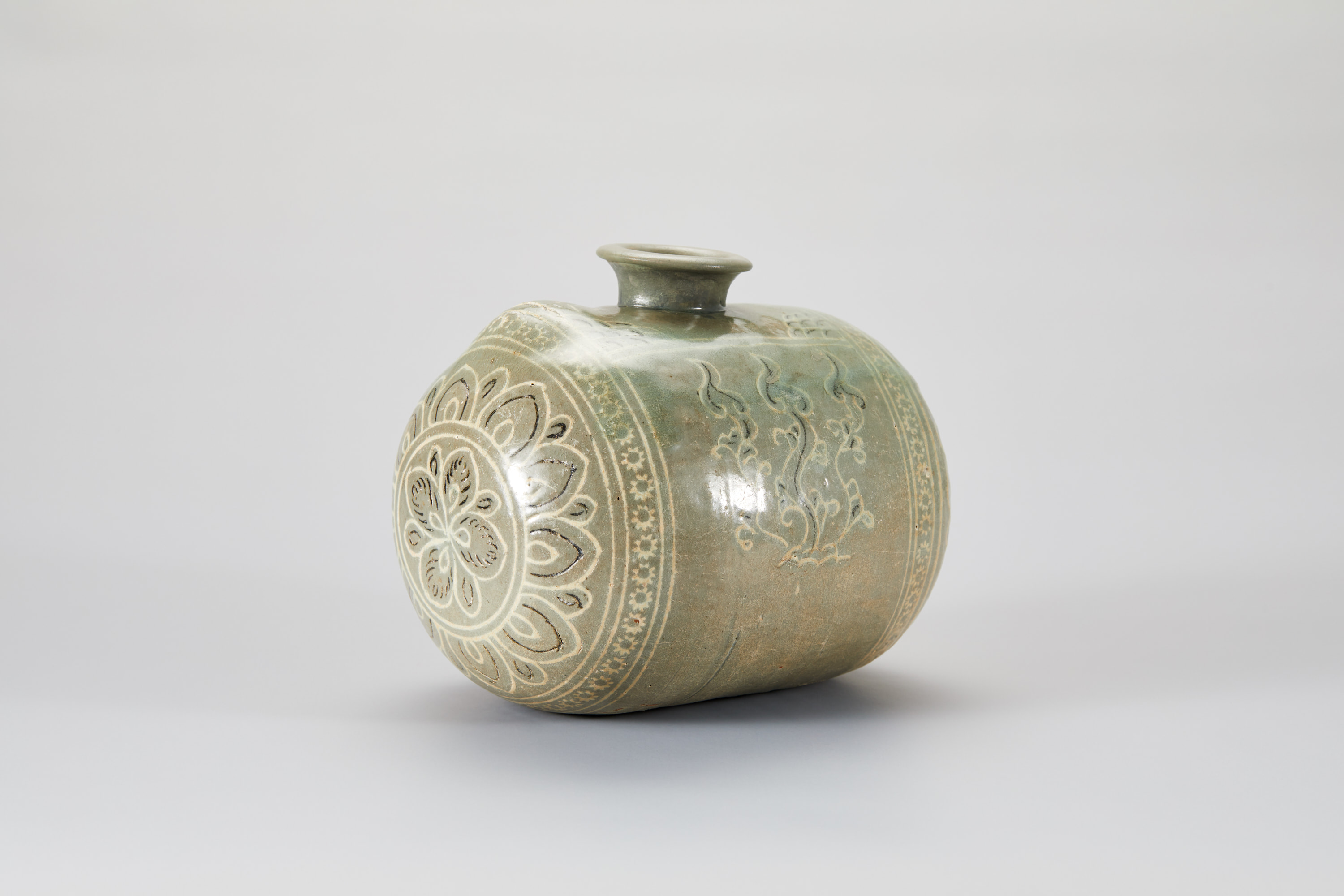 This cylindrical bottle has a flared opening at the center top. Geometric floral motifs adorn the side angled toward us, and the large central register features a looser flower motif. The bottle is light brown, the decorations are mostly thin white lines, and the top half has a light green glaze.