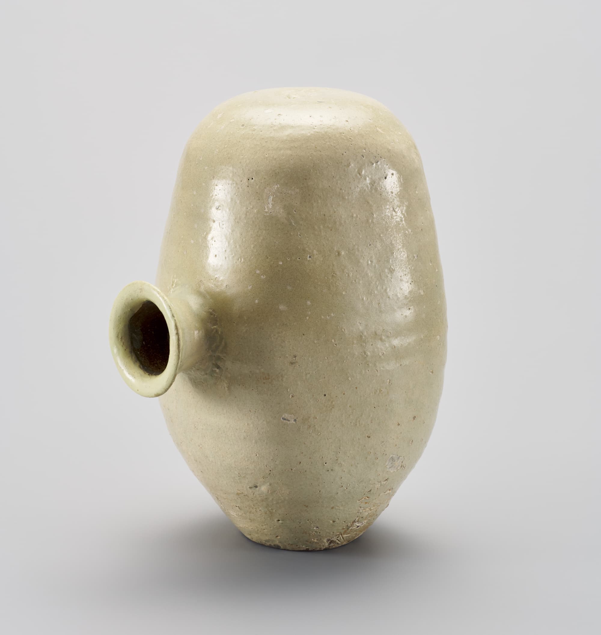This vertically oriented cream-colored vessel bulges slightly at center and rests on a flat bottom. At front left, in the center of the bulge, is a flared opening that protrudes from the body of the bottle.
